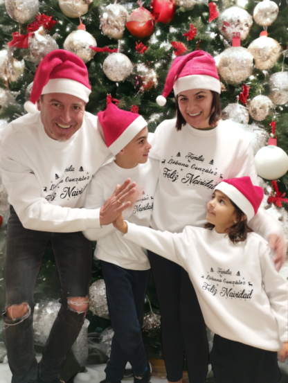 Padre, madre, hijo, hija, con sudaderas de navidad blancas y el apellido de la familia personalizado con un árbol de navidad de fondo, en el que el padre y la hija se chocan la mano.