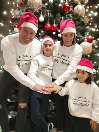 Padre, madre, hijo, hija, con sudaderas de navidad blancas y el apellido de la familia personalizado con un árbol de navidad de fondo, en la que los 4 juntan sus manos en el centro.