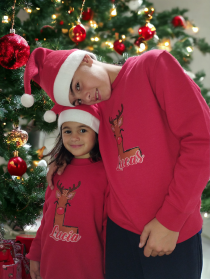 Un niño y una niña con sudadera de navidad roja con inicial con forma de reno y nombre personalizado con un árbol de navidad de fondo
